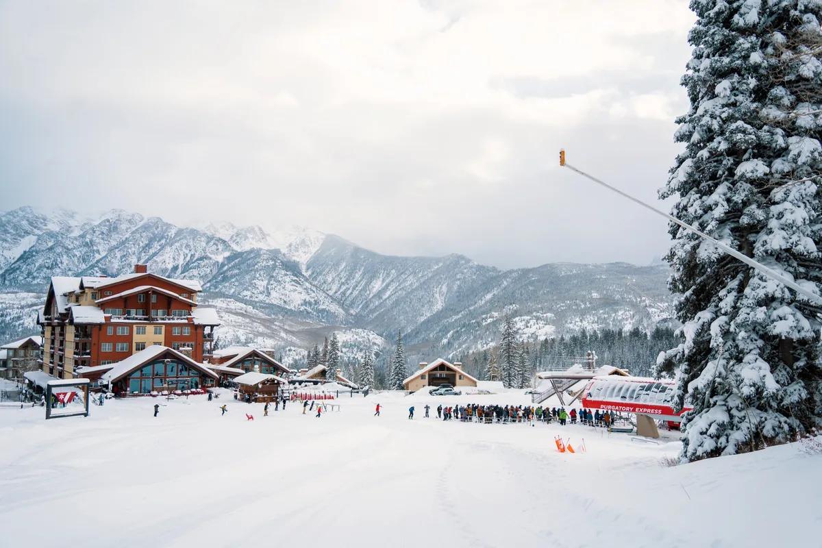 A lively winter scene capturing snow-covered mountains, skiers and snowboarders in action, resort buildings, and the nearby ski lift station. The image pulses with excitement and activity, with groups gathering at the base area and the stunning Needles Mountain Range visible in the background.