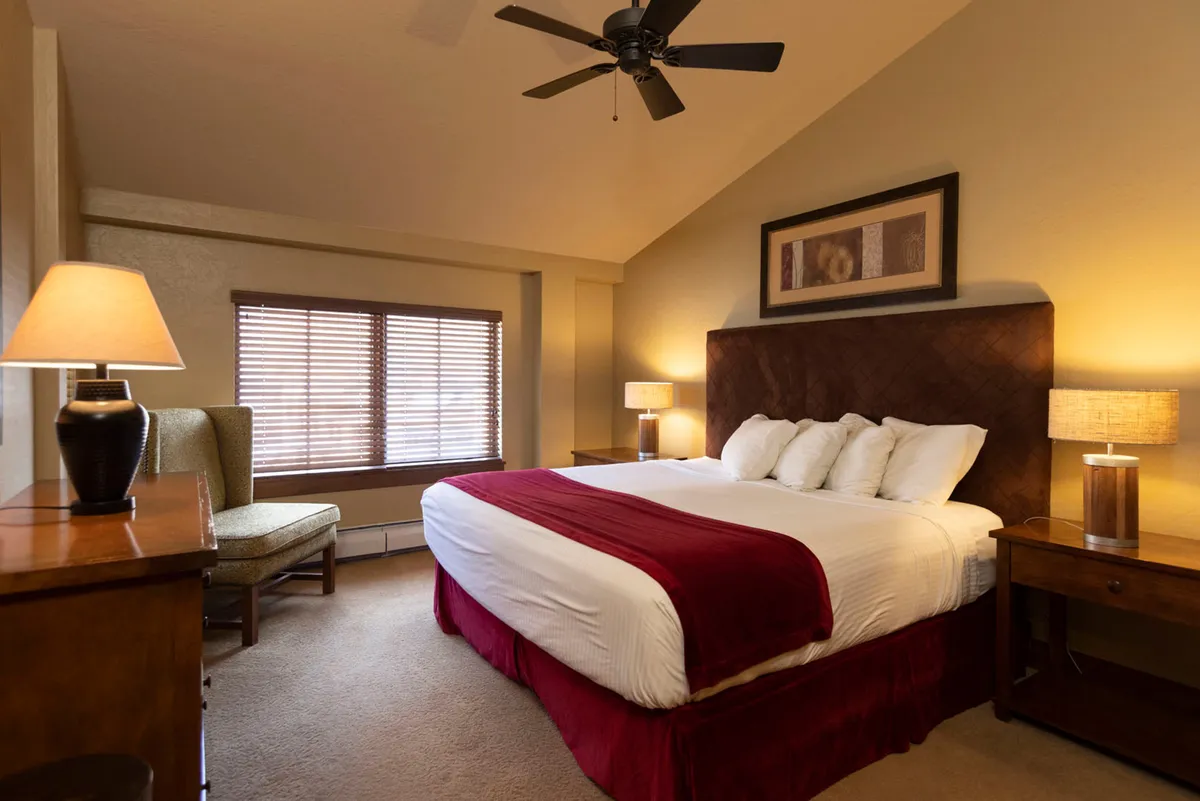 This image showcases a large, inviting bed with crisp white bedding and red accents, flanked by wooden nightstands and soft-glowing lamps. A plush chair sits by a sunlit window, emphasizing the restful and tranquil atmosphere of the bedroom.