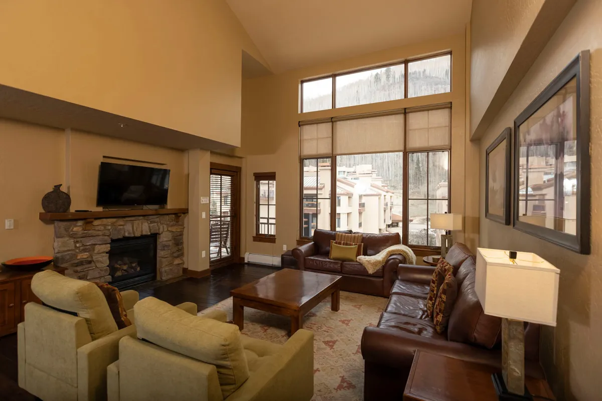 The image showcases a spacious, elegantly furnished living area featuring a grand stone fireplace as the centerpiece. Plush sofas are arranged for intimate gatherings, while expansive windows frame breathtaking mountain vistas just beyond. The golden glow of sunset casts a warm light across the room, inviting guests to relax and savor the serene alpine setting.