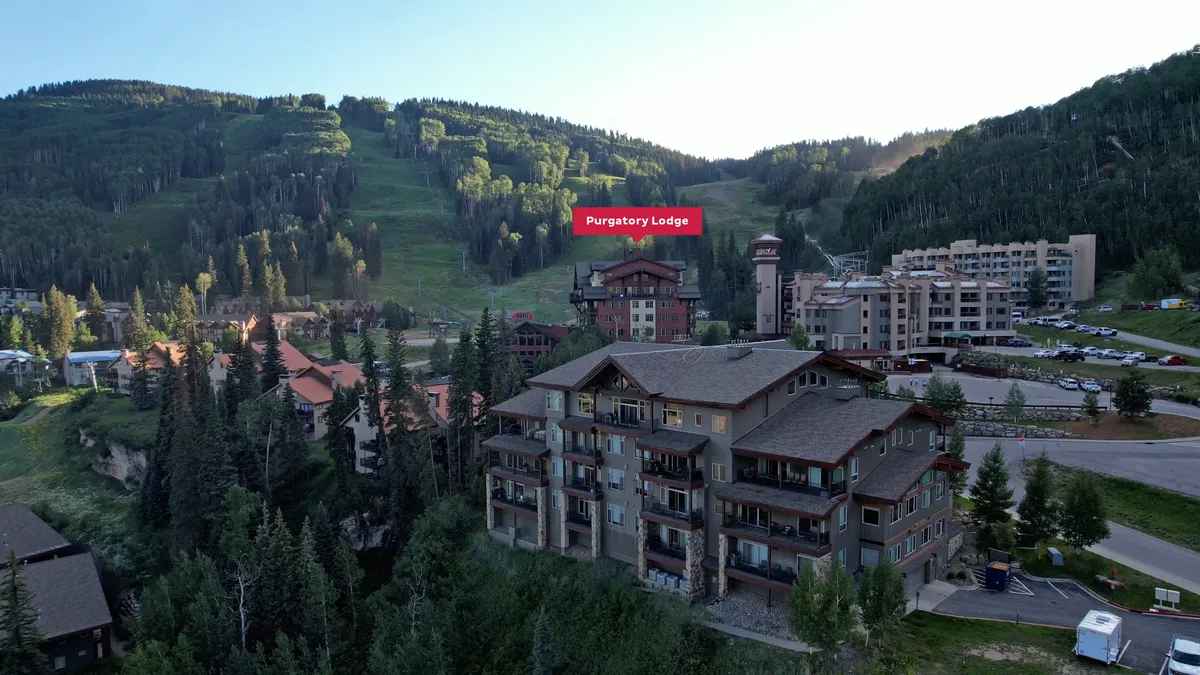 An exterior view of Purgatory Lodge set against a backdrop of majestic mountains and adjacent alpine buildings. The landscape radiates tranquility and adventure, with visible trails and vast open spaces ideal for all-season recreation.