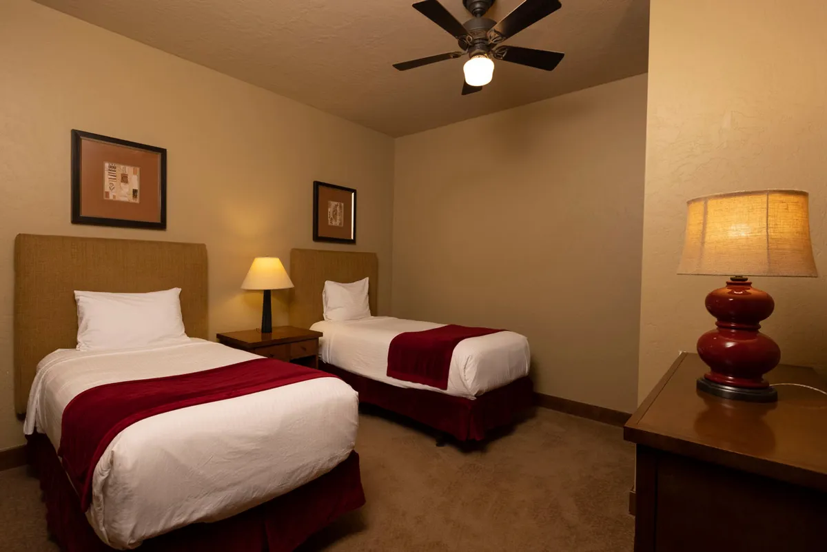A cozy bedroom featuring two twin beds topped with comfortable linens, separated by a table lamp, and accented by framed artwork. The setting feels intimate and perfect for families or children.