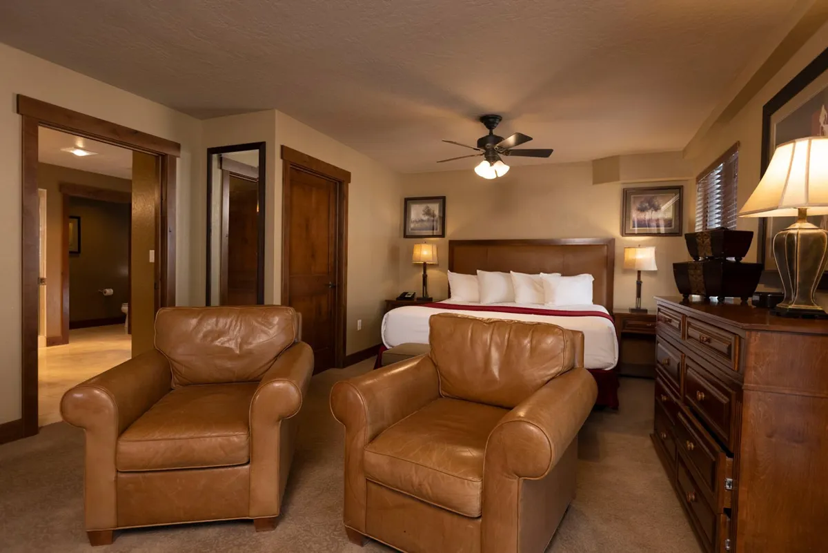 A welcoming queen bedroom scene, highlighted by a queen bed with fresh white bedding, two leather armchairs perfect for reading or relaxing, a wooden dresser, and a softly-lit table lamp creating a warm atmosphere.