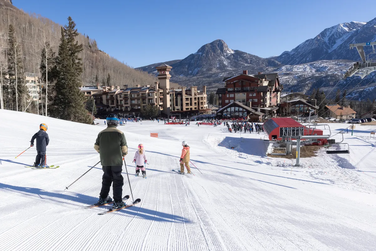 A dynamic image with skiers navigating the slopes against a dramatic mountain backdrop, flanked by resort buildings and a visible ski lift. Families and children are seen enjoying their first ski lessons, while others make their way to après-ski relaxation.