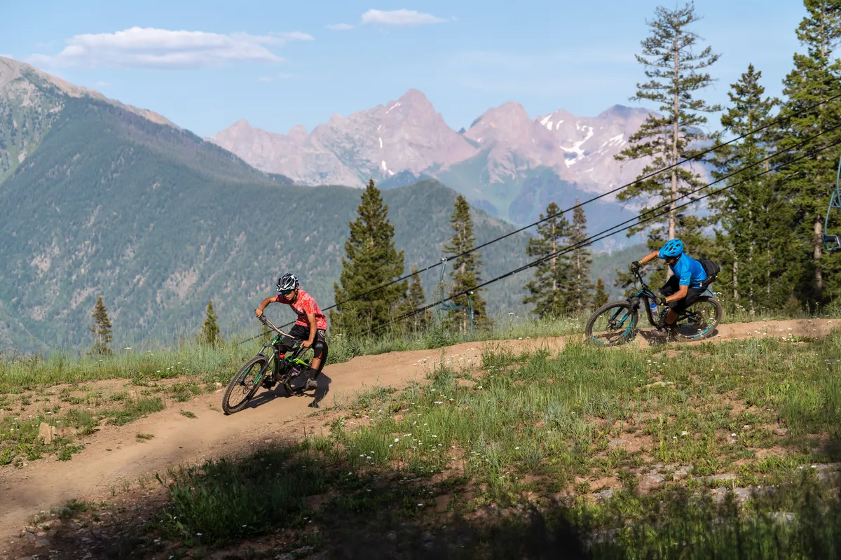 The image shows a group of mountain bikers traversing a rugged mountain landscape filled with lush trees and dramatic slopes. The expansive natural scenery and active bikers capture the sense of adventure and exploration. Sunlight filters through the trees, highlighting the energetic mood.