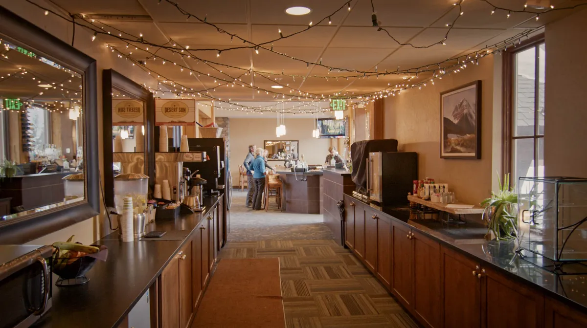 A welcoming lounge area featuring string lights overhead, a cozy seating arrangement, a coffee station, a bar area, and captivating mountain-themed artwork. The space invites relaxation, conversation, and entertainment, with a TV visible in the background and plush chairs arranged for group gatherings.
