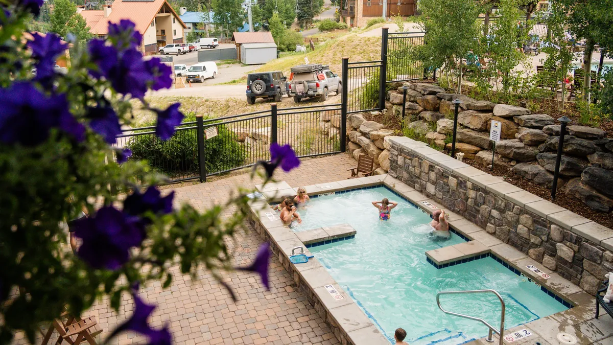 This exterior image showcases the inviting swimming pool surrounded by lush flowers, with people enjoying the water and sunbathing. The pool area is lively yet relaxing, reflecting a resort-style recreation atmosphere.